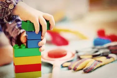 Kinderhände die Legosteine aufeinanderstapeln, daneben liegen Spielsachen auf dem Boden